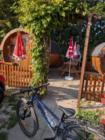 Schlaffässer mit Sonnenschirmen und Grillplatz, davor ein Fahrrad